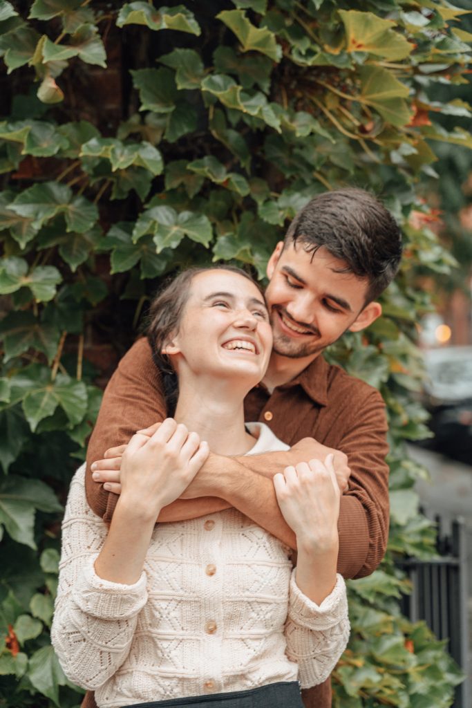 Trennis embracing Maria from behind beneath climbing greenery